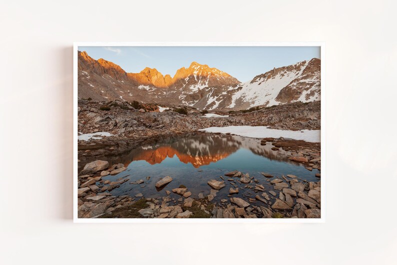 May include: A framed landscape photograph captures a mountain range at sunrise. The peaks are illuminated with golden light, reflected in a still pool of water. The foreground is filled with rocks and the sky is a soft blue.