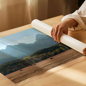 May include: A landscape print featuring a mountain range, a field with horses, and a forest. The print is being unrolled on a wooden surface. The color palette includes shades of blue, green, brown, and yellow.