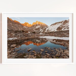 May include: A framed landscape photograph of a mountain range at sunrise. The peaks are illuminated with golden light, reflecting in a still lake. Snow patches and rocky terrain are visible.