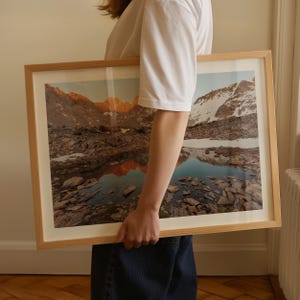 May include: A framed landscape print featuring a mountain range reflected in a lake. The artwork has a light wood frame and showcases a scenic view with snow-capped peaks, rocky terrain, and a serene body of water.