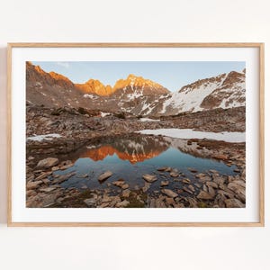 May include: A framed landscape photograph captures a mountain range at sunrise. The peaks are illuminated with golden light, reflecting in a still lake. Snow patches dot the mountainsides, and the foreground is filled with rocks.