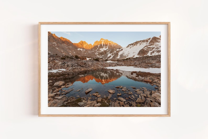 May include: A framed landscape photograph captures a mountain range at sunrise. The peaks are illuminated with golden light, reflecting in a still lake. Snow patches dot the mountainsides, and the foreground is filled with rocks.