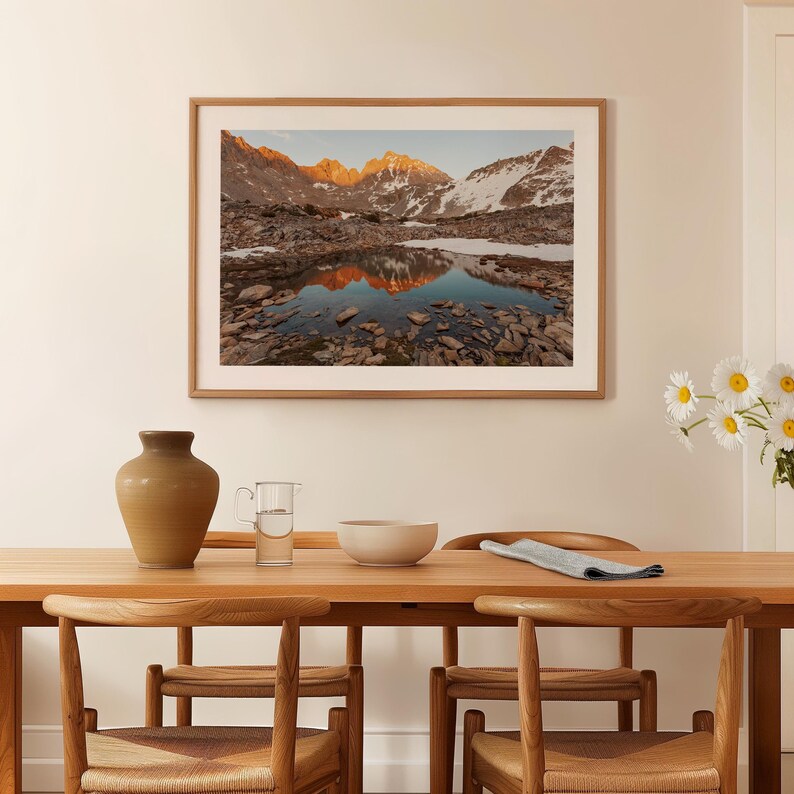 May include: A framed landscape photograph depicts a mountain range with snow-capped peaks reflected in a still lake. The image is in a light brown wooden frame, hanging above a wooden dining table with chairs, a vase, and a bowl.