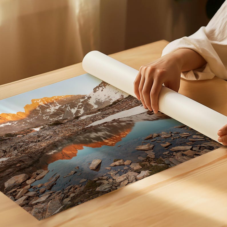 May include: A landscape print featuring a mountain range reflected in a lake. The image has a warm color palette with golden hues on the mountain peaks and a clear blue lake. The print is being unrolled on a wooden surface.