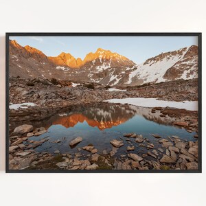 May include: A framed landscape photograph featuring a mountain range with snow-capped peaks and a reflective lake. The mountains are illuminated by the golden light of sunrise or sunset, casting a warm glow. The lake reflects the mountains and sky.