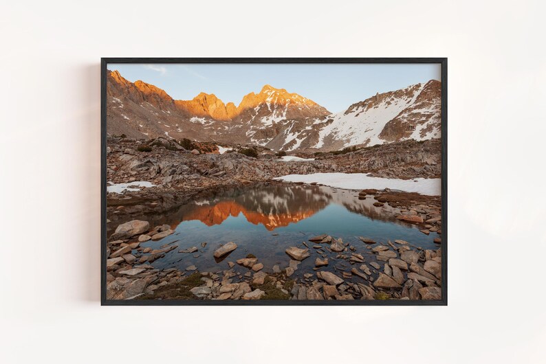 May include: A framed landscape photograph featuring a mountain range with snow-capped peaks and a reflective lake. The mountains are illuminated by the golden light of sunrise or sunset, casting a warm glow. The lake reflects the mountains and sky.