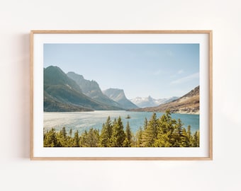 May include: A framed landscape print featuring a serene mountain lake scene. The image showcases a clear blue sky, a lake surrounded by mountains, and evergreen trees in the foreground. The print is encased in a light brown wooden frame.