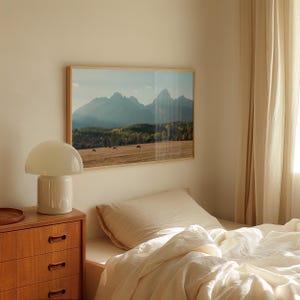 May include: A framed landscape photograph of a mountain range with a field and trees in the foreground. A white mushroom-shaped lamp sits on a wooden dresser. A bed with cream-colored bedding is in the foreground.