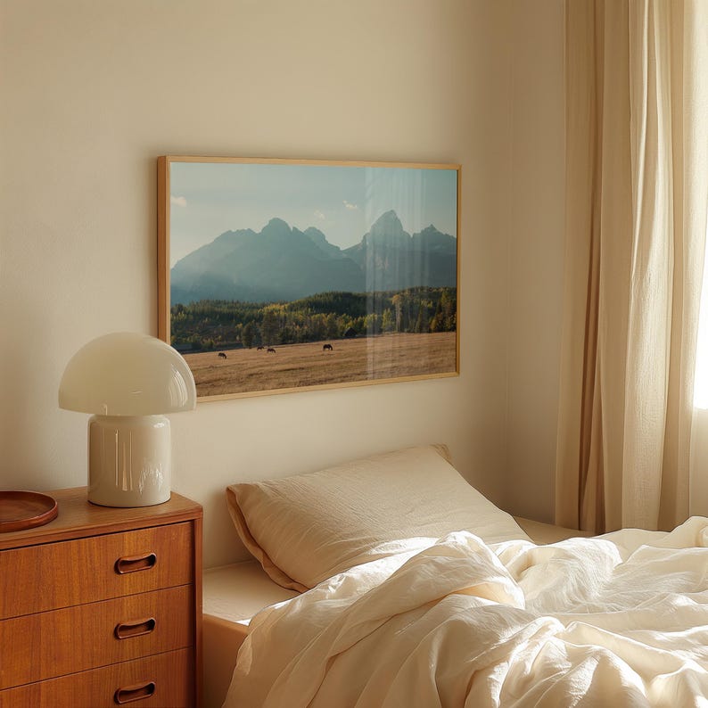 May include: A framed landscape photograph of a mountain range with a field and trees in the foreground. A white mushroom-shaped lamp sits on a wooden dresser. A bed with cream-colored bedding is in the foreground.