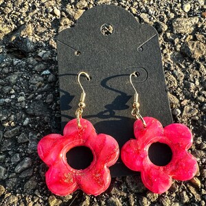 May include: A pair of pink flower-shaped earrings with gold hooks. The earrings have a glittery finish and a hole in the center.