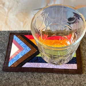 May include: A clear, patterned glass tumbler sits on a colorful, patchwork fabric coaster. The coaster features a geometric design with various fabric strips in shades of blue, red, orange, and purple, framed by a dark brown border. The tumbler is empty.