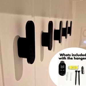 Könnte beinhalten: Reihe schwarzer Wandhaken, die auf einer weißen Oberfläche montiert sind. Die Haken haben ein abgerundetes, T-förmiges Design. Das Bild zeigt auch die Hardware, die im Lieferumfang der Haken enthalten ist, einschließlich Schrauben und einer Wasserwaage.
