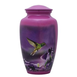 May include: A cylindrical cremation urn with a smooth, glossy finish in shades of magenta and deep purple. The body of the urn features a printed graphic of a green and yellow hummingbird in flight, hovering near dark purple iris flowers.