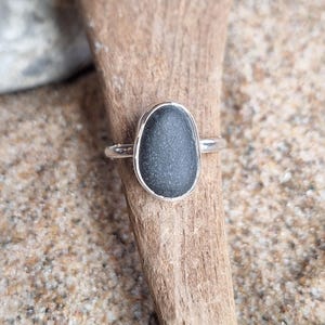 May include: A silver ring featuring a dark gray, oval-shaped stone. The stone is set in a simple bezel setting, and the band is thin and polished. The ring is displayed on a piece of driftwood against a sandy background.