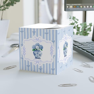 May include: A square notepad holder with a blue and white striped pattern. The design includes framed illustrations of blue hydrangeas in a blue and white vase. Decorative bows are scattered across the surface.