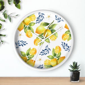 Può includere: Un orologio da parete rotondo con cornice bianca e quadrante bianco caratterizzato da un motivo ad acquerello di limoni gialli, foglie verdi, piccoli fiori bianchi e rametti blu scuro. L'orologio ha tre sottili lancette bianche.