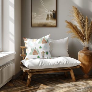 Può includere: Un cuscino bianco con un motivo ad acquerello di una baita, sci, fiocchi di neve e alberi. Il cuscino è su una panca di legno con un cuscino bianco dietro. Un quadro incorniciato e un vaso con piante secche sono sullo sfondo.
