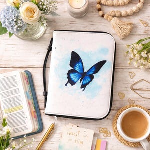 May include: White Bible cover with a blue butterfly design. The butterfly has detailed wing patterns and is set against a light blue watercolor background. The cover has a black handle and trim. A gold pen and a card with the text "Trust in Him" are also visible.