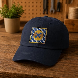 Puede incluir: Una gorra de béisbol azul marino con un parche cuadrado en la parte delantera. El parche muestra un fondo de patrón de cuadros azul y blanco con una forma de sol amarilla en el centro, que contiene dos llaves inglesas azules cruzadas.
