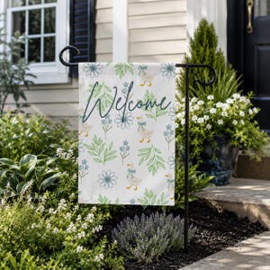 May include: A rectangular garden flag featuring a white background with a repeating pattern of light green leaves, blue flowers, and white ducks wearing blue bows. The word Welcome is written in a dark blue cursive script across the center.