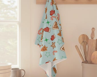 May include: A light blue kitchen towel featuring a pattern of gingerbread biscuits, candy canes, and red mittens. The towel is hanging on a wooden rack. The towel has a white interior and a light blue exterior.