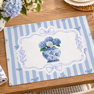 May include: A rectangular placemat featuring a light blue and white vertical striped pattern with small blue bows. The centre displays a white scalloped frame containing an illustration of a blue and white patterned vase filled with blue hydrangeas.