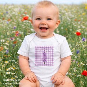 May include: A smiling baby wearing a white onesie with a purple gingham design featuring a purple flower and a bee. The onesie has a square design with a bow at the top. The baby is sitting in a field of wildflowers.