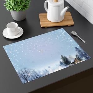 May include: A rectangular placemat with a winter scene. The placemat features a light blue background with falling snowflakes and a watercolor image of a snowy forest and a cabin. A white teacup and saucer are on the left.