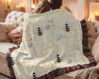 Puede incluir: Manta crema con un estampado de abejas y panales. La manta presenta abejas negras, amarillas y blancas, junto con formas de panal y detalles florales. La manta tiene un borde con flecos con hilos rojos, negros y amarillos.