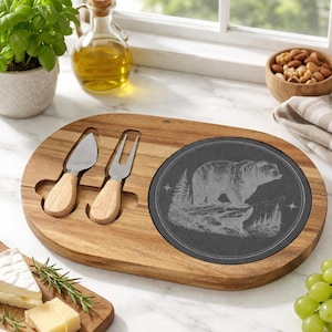 May include: Wooden cheese board with a dark gray slate insert featuring a bear design. Includes two cheese knives with wooden handles. The board is oval-shaped and has cutouts for the knives.