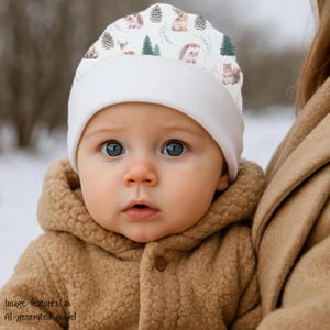 Op de afbeelding: Een witte baby beanie met een bosdierenpatroon, met eekhoorns, herten, egels en dennenbomen. De beanie heeft een omgeslagen rand en wordt gedragen door een baby met blauwe ogen en een bruine jas met capuchon.