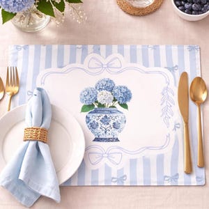 Puede incluir: Un mantel individual a rayas azules y blancas con un diseño floral. El mantel individual presenta un jarrón con hortensias azules y un borde decorativo con lazos. También se ven cubiertos dorados, un plato blanco y una servilleta azul.