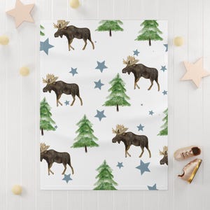 Puede incluir: Manta de beb&eacute; blanca con un estampado repetido de alces marrones, pinos verdes y estrellas azules. La manta est&aacute; hecha de material suave, perfecta para una guarder&iacute;a o como regalo de baby shower.