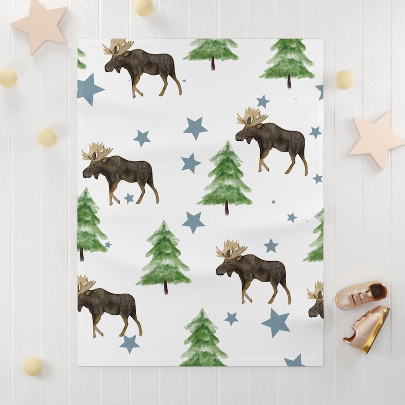 Puede incluir: Manta de beb&eacute; blanca con un estampado repetido de alces marrones, pinos verdes y estrellas azules. La manta est&aacute; hecha de material suave, perfecta para una guarder&iacute;a o como regalo de baby shower.