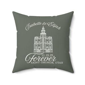 May include: A square, gray pillow with white text and a detailed illustration of a building. The text reads "Isabelle & Elijah", "11.15.25", and "Forever Saint George, Utah".