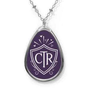 May include: Silver-toned necklace with a purple oval pendant. The pendant features a white shield design with the letters "CTR" inside, surrounded by hearts and decorative flourishes. The chain has small, round beads.