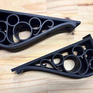 May include: Two black decorative shelf brackets with a repeating circular and curved design. The brackets are triangular in shape and appear to be made of a solid material. The brackets are shown on a light brown wooden surface.