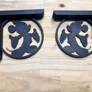 May include: Two black decorative shelf brackets. Each bracket features a silhouette of a famous cartoon character within a circular frame. The brackets are designed to be mounted on a wall and provide support for shelves.