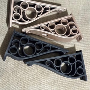 May include: Four decorative shelf brackets in shades of beige and black. Each bracket features a stylized Mickey Mouse head design with curved lines and circular cutouts. The brackets are made of a solid material and are designed for wall mounting.