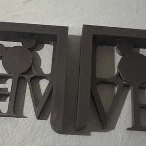 May include: Two brown metal shelf brackets. Each bracket is shaped with the word "LOVE" and a Mickey Mouse head silhouette. The brackets are designed to be mounted on a wall for decorative shelving.