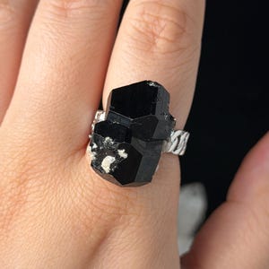 Puede incluir: Un anillo de turmalina negra con una gema en bruto sin cortar. El anillo tiene una banda plateada con un diseño texturizado. La gema es de color negro intenso con algunas inclusiones blancas y grises. El anillo se muestra en una mano.