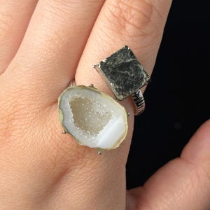 Puede incluir: Un anillo de plata con dos engastes de piedra únicos. Uno tiene una piedra cuadrada de color gris oscuro, mientras que el otro muestra una rodaja de geoda con un interior blanco y un borde verde claro. Es una pieza de joyería.