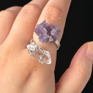 Peut inclure: Une bague ouverte de couleur argentée ornée de deux pierres distinctes : un amas de petits cristaux violets texturés sur le dessus et un cristal transparent, allongé et pointu sur le dessous, montés sur un anneau métallique strié.