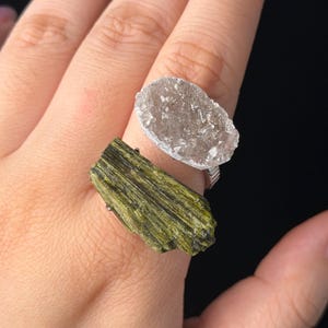Op de afbeelding: Een ring met twee stenen, bestaande uit een ruw, olijfgroen toermalijnkristal met verticale strepen en een ronde, doorschijnende druzy kwartssteen met een sprankelend, kristallijn oppervlak, gemonteerd op een metalen band.