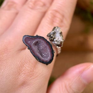 May include: A silver ring with a geode design. The ring features a large, open geode stone with a dark purple interior and a smaller, textured stone. The geode has a dark border. The ring is a unique piece of jewellery.