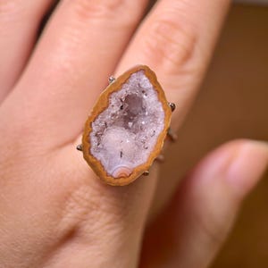 May include: A unique ring featuring a geode stone with a brown and white outer edge and a crystal-filled centre. The ring's setting is a silver-toned metal. The geode measures approximately 3.8 cm in length.