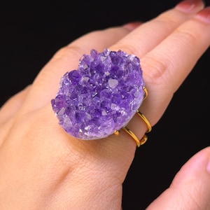 Op de afbeelding: Een ring met een grote, ruwe, dieppaarse amethist druzy steen op een dunne, gepolijste goudkleurige metalen band. Het amethistcluster vertoont talloze kleine, grillige kristalpunten die het licht vangen en een textuur creëren.