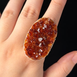 May include: A ring featuring a large, oval-shaped, deep amber-coloured crystal cluster with a textured, faceted surface. The crystal is set on a simple band, showcasing a raw, natural appearance with rich, warm orange and brown tones throughout.