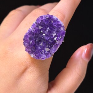 May include: A large oval ring featuring a cluster of deep purple amethyst druzy crystals. The raw, textured gemstone surface displays multifaceted points that catch the light, set securely onto a simple metallic band worn on a finger.