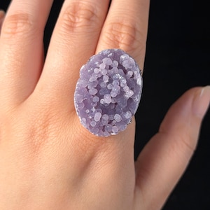 May include: A statement ring featuring a large, oval-shaped purple druzy gemstone with a textured, bubbly surface. The stone is set in a metallic frame that follows the irregular, organic contour of the crystal cluster, creating a bold look.